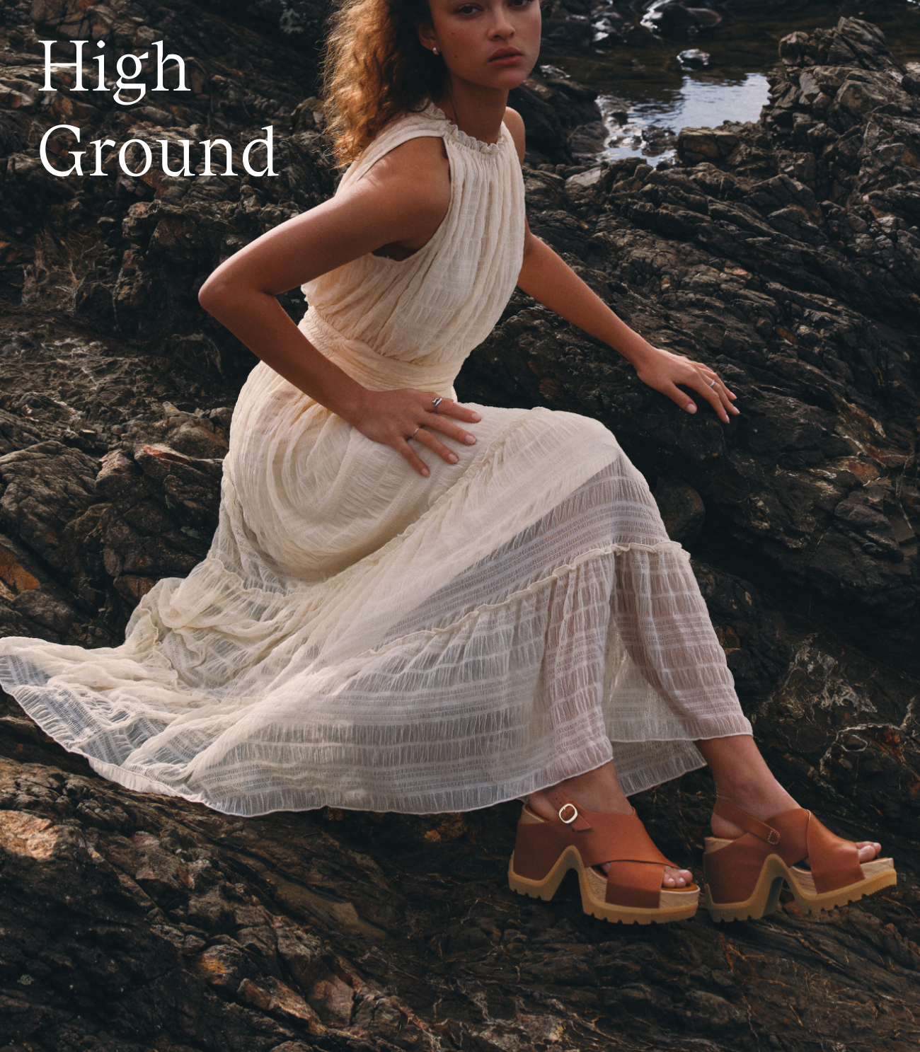 Woman in white dress sitting atop dark rock wearing ROAM'N RISE Sandals